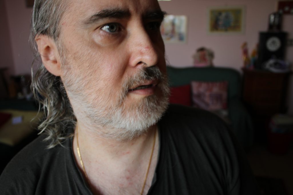 Primo piano di Marco De Luca con barba e capelli grigi che guarda di lato in un ambiente domestico sfocato sullo sfondo.